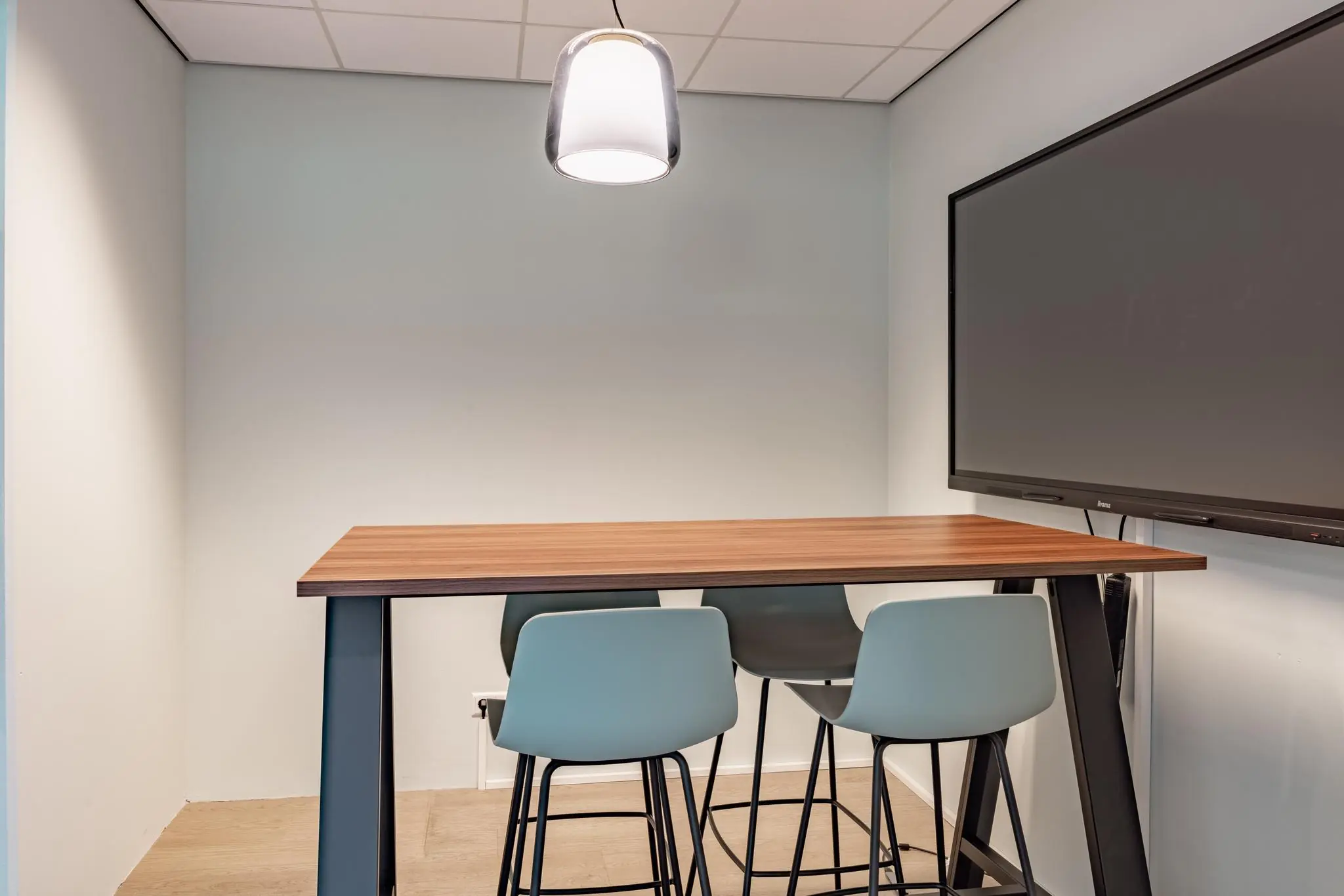 Moderne vergaderruimte met hoge tafel, vier barkrukken, hanglamp en een groot digitaal scherm aan de muur.